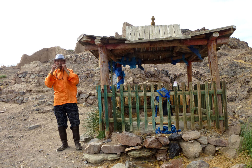 Ein Mann aus der Mongolei steht neben einer Tempelanlage. Er trägt einen Hut und spielt auf einer Maultrommel